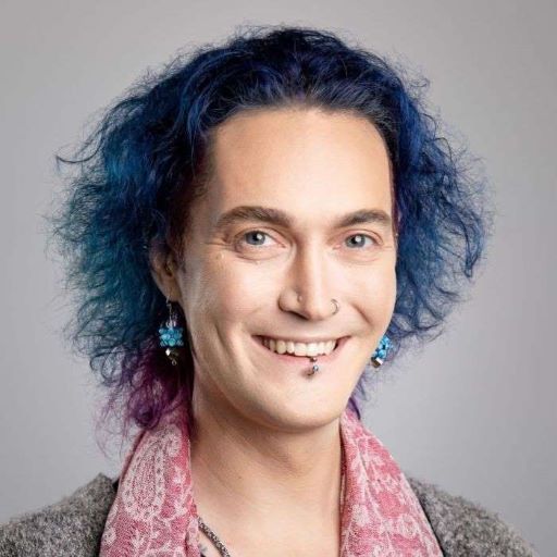 Headshot: Jewel, a white person with blue-dyed hair cut into a muppetish bob, is wearing a watermelon-colored pashmina scarf, blue glass teardrop earrings shaped like a blobby star, and a lip ring in the middle of her smile.